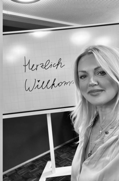 HR Seminare und Business Coaching About Work GmbH Frau mit blonden Haaren steht neben einer Tafel mit der Aufschrift "Herzlich Willkommen".
